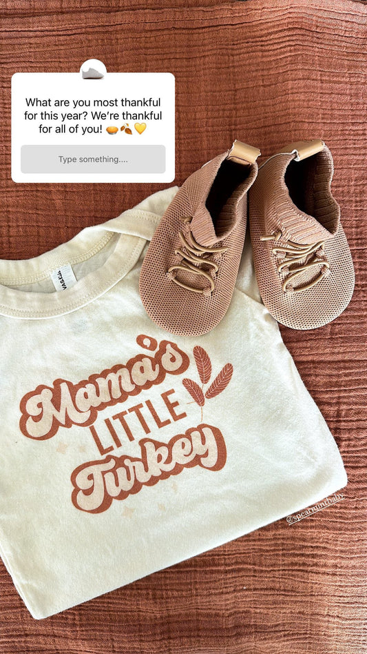 Flat lay of tan knit HARTS Bootees barefoot boots beside a cream “Mama’s Little Turkey” toddler tee on a rust-colored fabric — cozy Thanksgiving outfit for babies and toddlers.