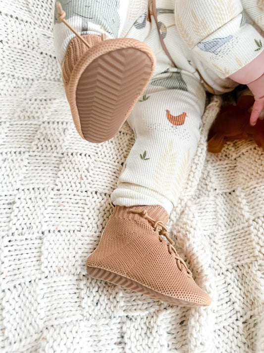 Close-up of a baby wearing tan knit HARTS Bootees barefoot boots while resting on a cream knit blanket — flexible, soft-soled shoes designed for natural movement.