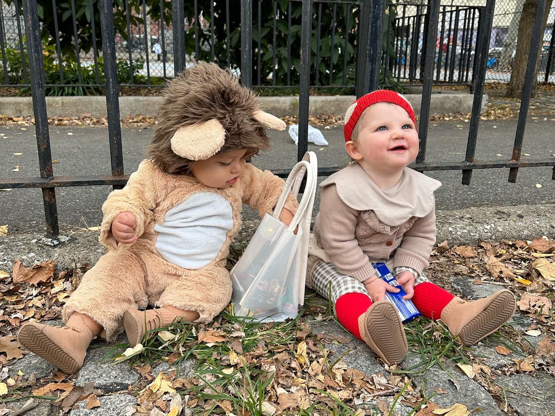 Two toddlers sitting outdoors in fall leaves, one in a lion costume and one in red tights, wearing HARTS Bootees barefoot boots and sharing Halloween treats.