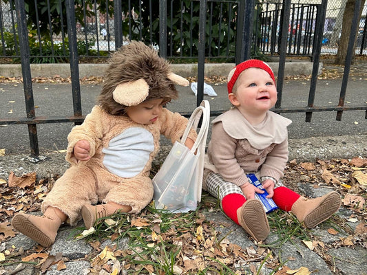 Two toddlers sitting outdoors in fall leaves, one in a lion costume and one in red tights, wearing HARTS Bootees barefoot boots and sharing Halloween treats.