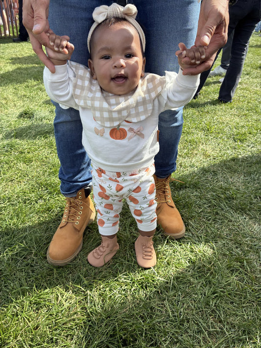 Baby in a fall pumpkin outfit wearing tan HARTS Park Boss boots, standing on grass holding a parent's hands